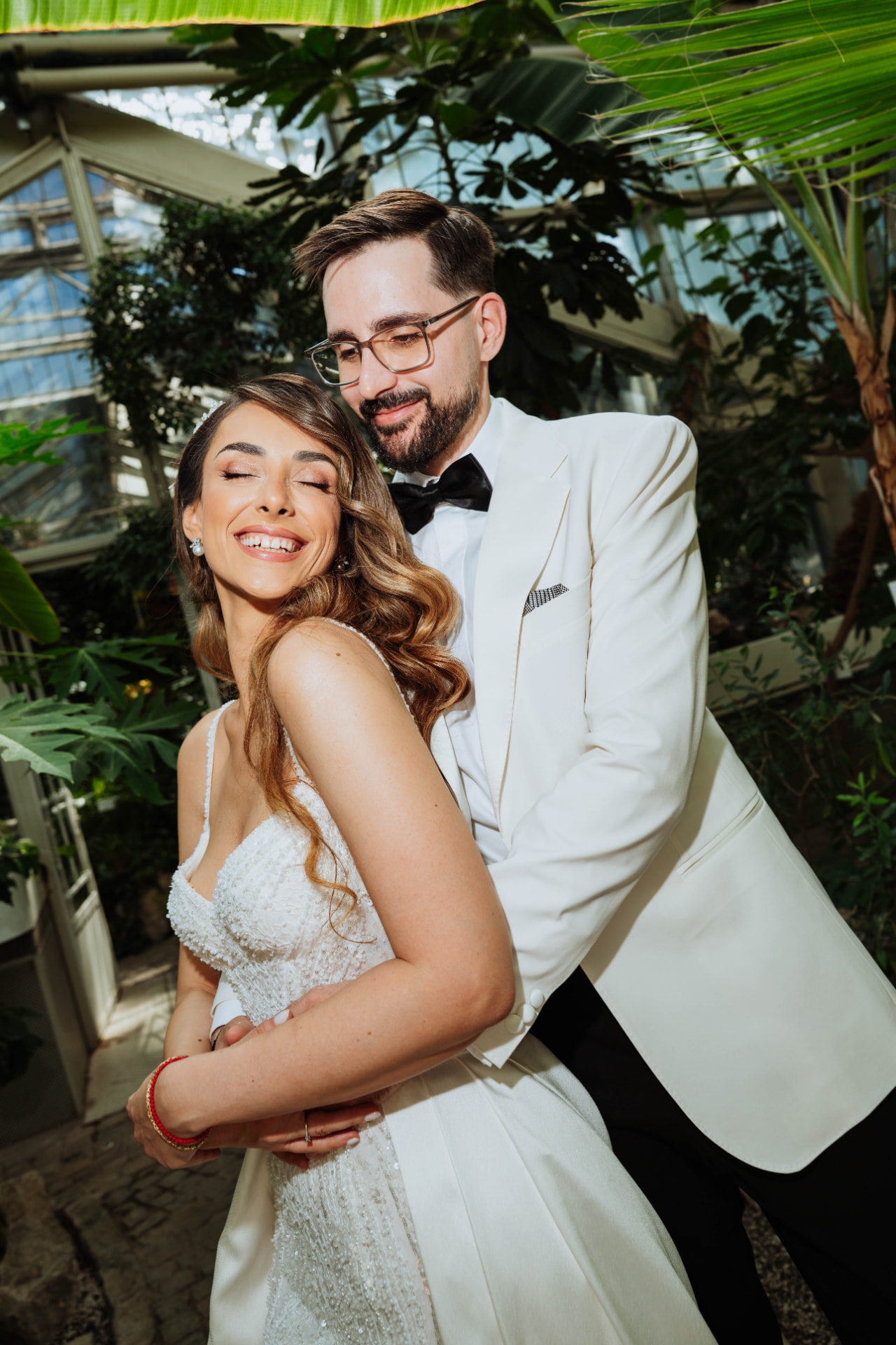 Groom in a white dinner jacket holding his bride from behind as she smiles with closed eyes inside a glasshouse full of palms.