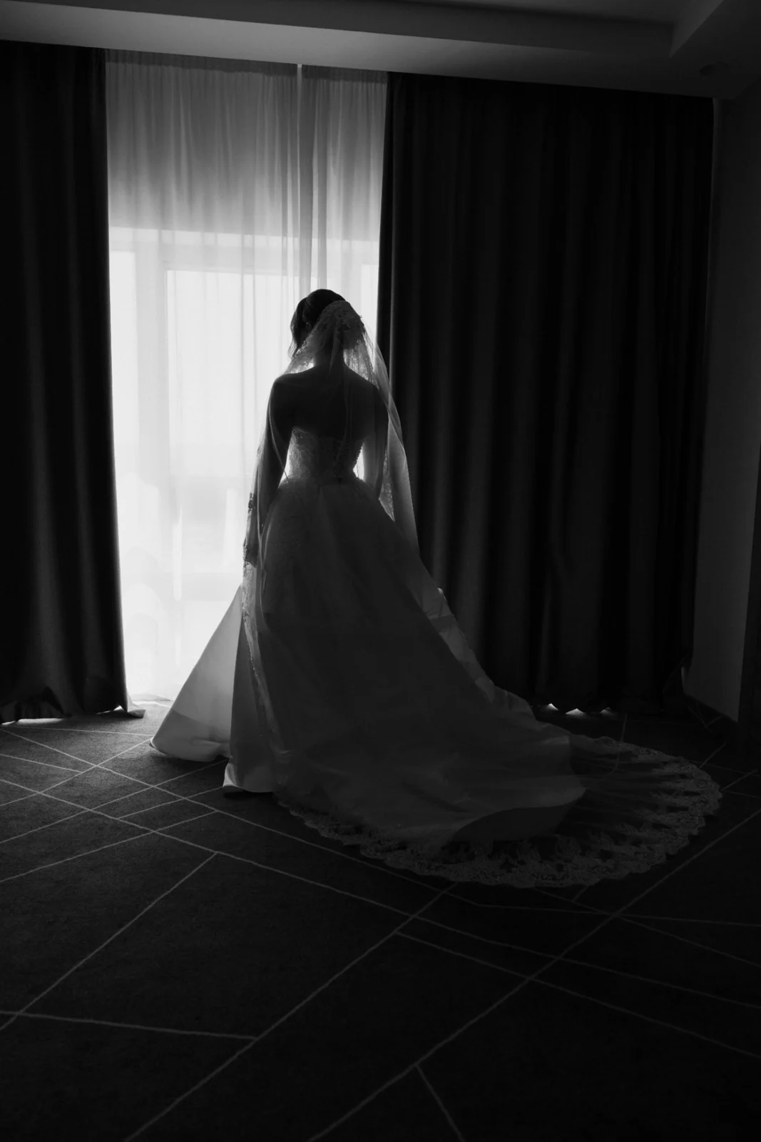 Black and white wedding photograph of a bride standing by a window in her wedding dress, photographed by Nemanja Potkonjak.