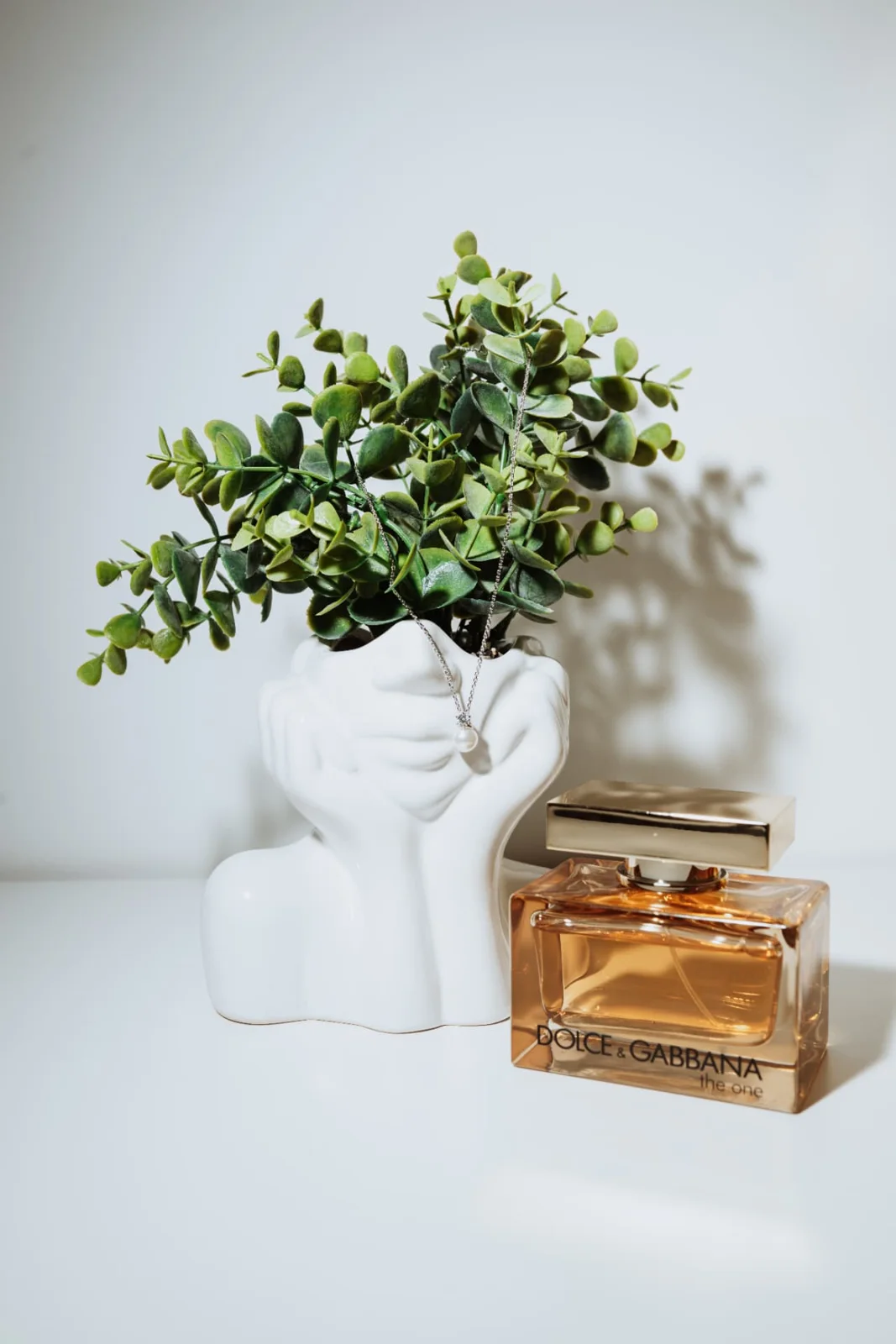 Bridal details: a white ceramic vase of eucalyptus with a pearl pendant necklace draped over it, beside a bottle of perfume.