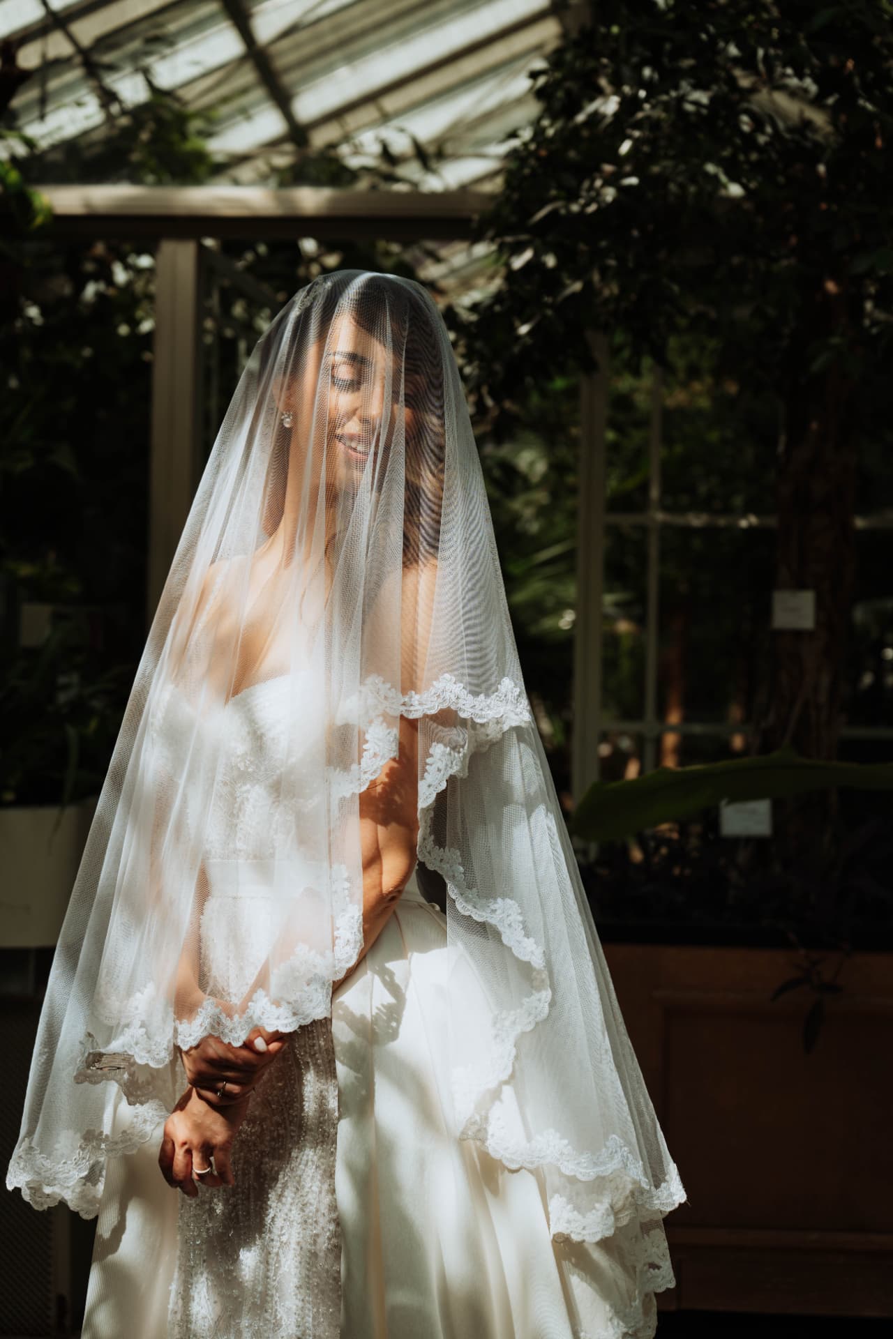 Bride standing with her eyes lowered beneath a long lace-edged veil in a greenhouse, soft daylight through the glass roof.